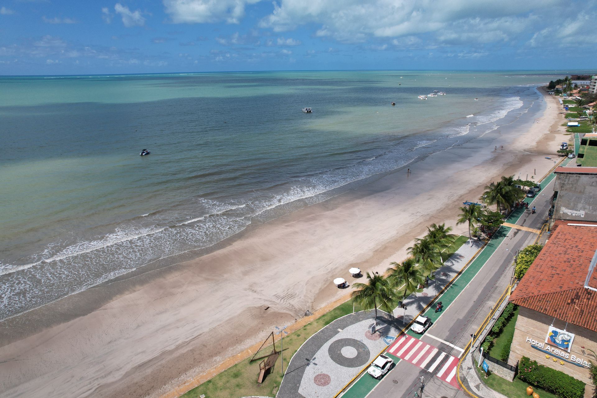 Praia costeira e calçadão junto a águas calmas azul-esverdeadas, com estrada, palmeiras e edifícios com telhados vermelhos.
