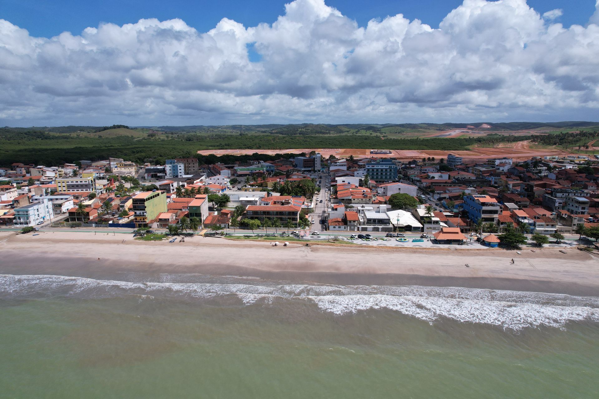 Vista aérea de uma cidade costeira com praia, ondas e edifícios coloridos sob um céu nublado.
