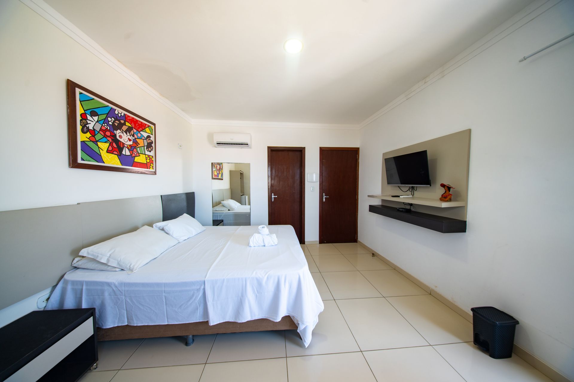 Quarto de hotel luminoso com cama, TV, obras de arte na parede e piso de azulejo branco.