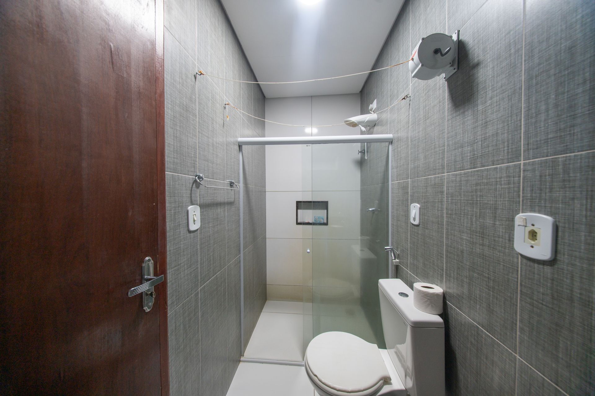 Pequeno banheiro revestido com azulejos, com vaso sanitário, chuveiro, porta de madeira e paredes cinza.