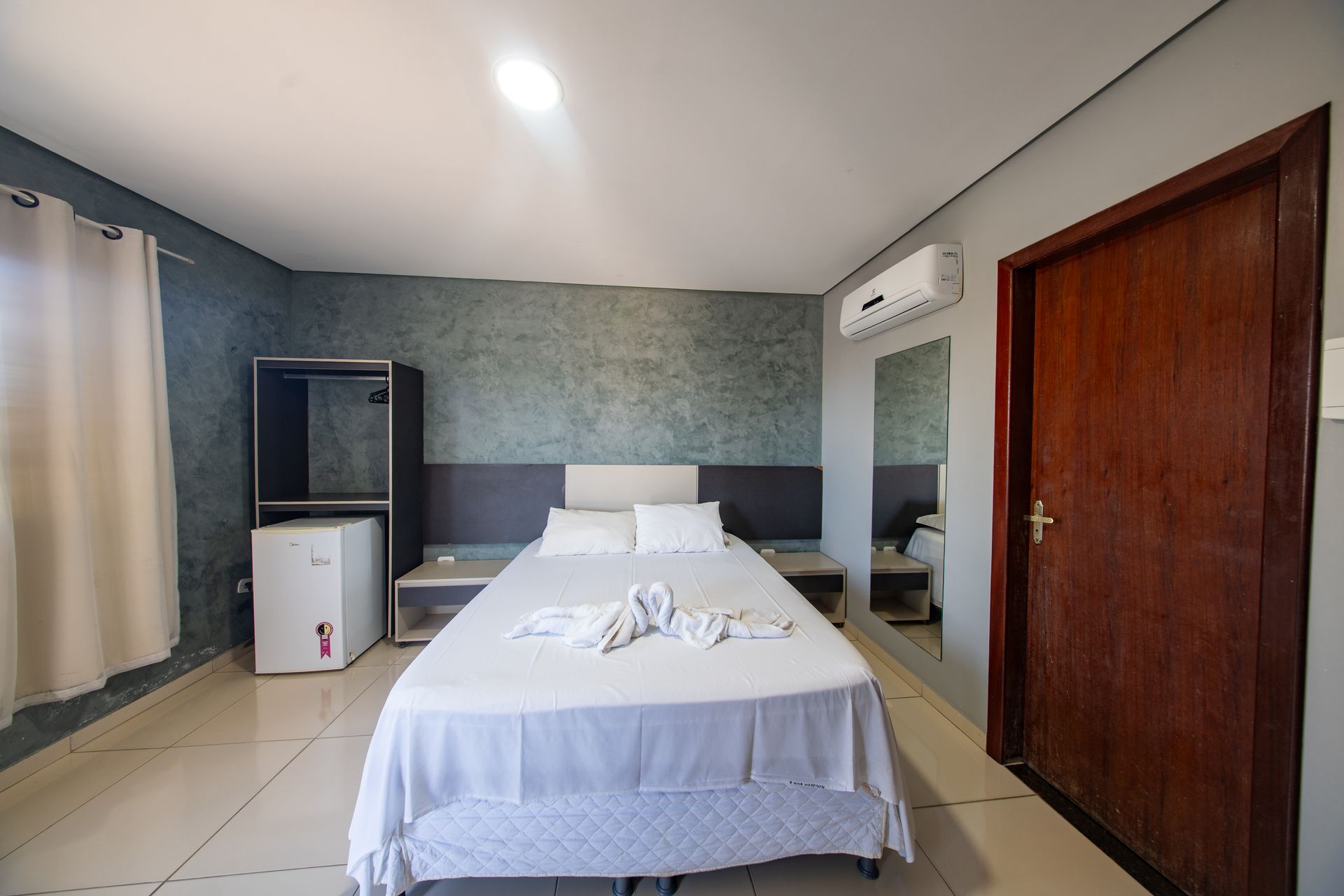 Quarto de hotel minimalista com uma cama de solteiro, armários brancos, espelho, ar condicionado e porta de madeira.