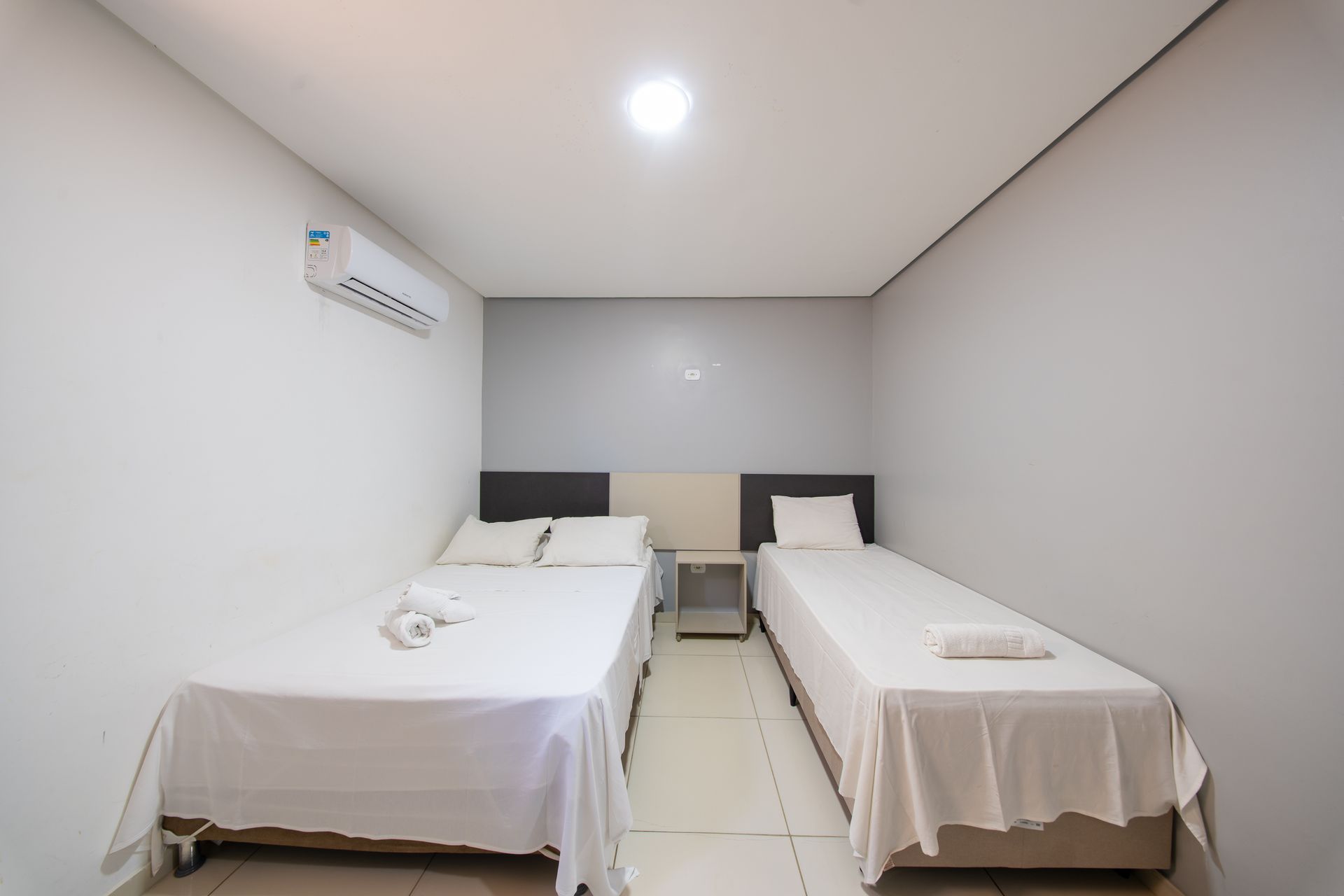 Quarto de hotel minimalista com duas camas de solteiro, lençóis brancos e uma pequena mesa de cabeceira.