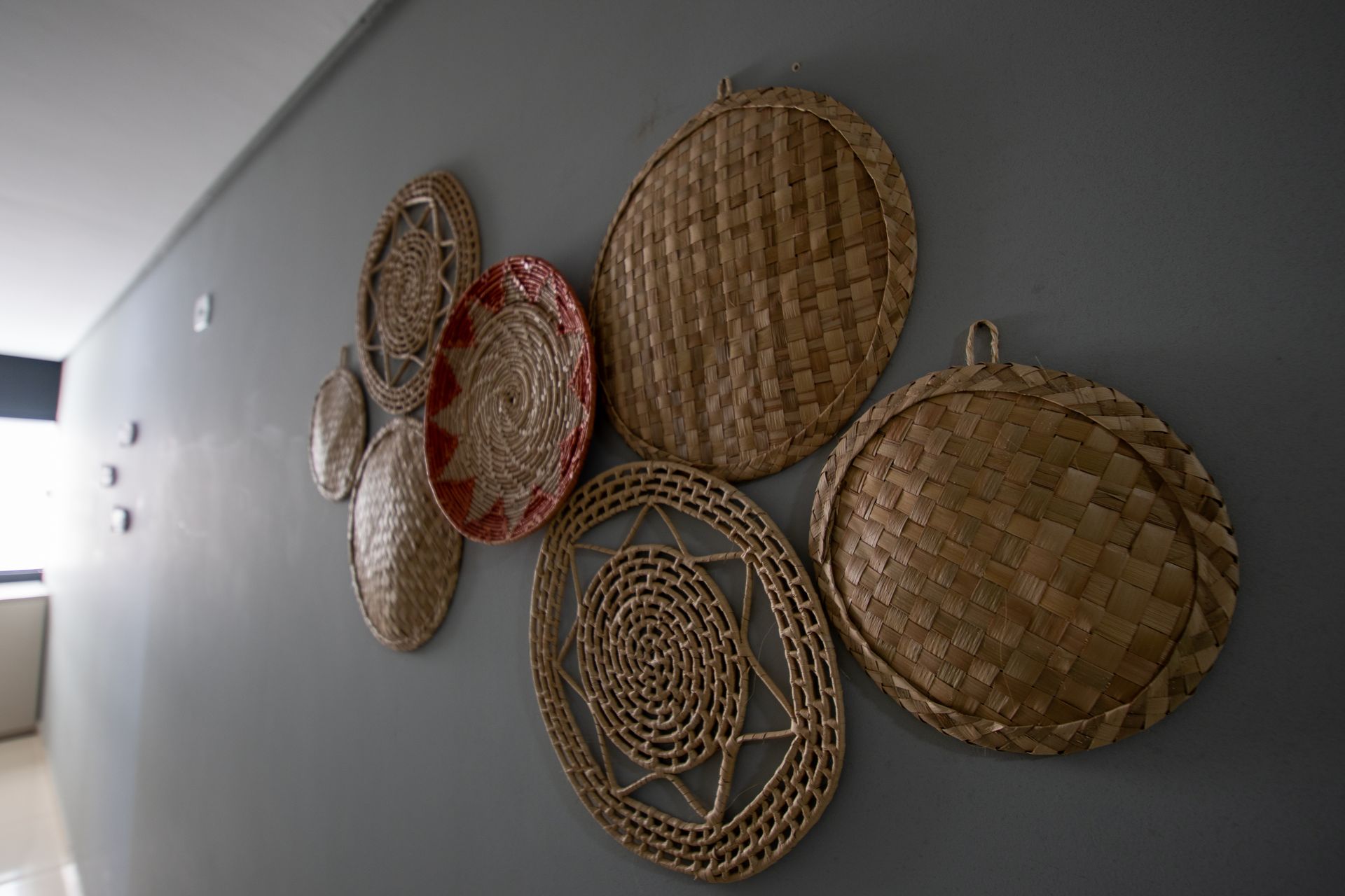 Cestos e bandejas de vime trançado dispostos de forma decorativa em uma parede cinza em um corredor.