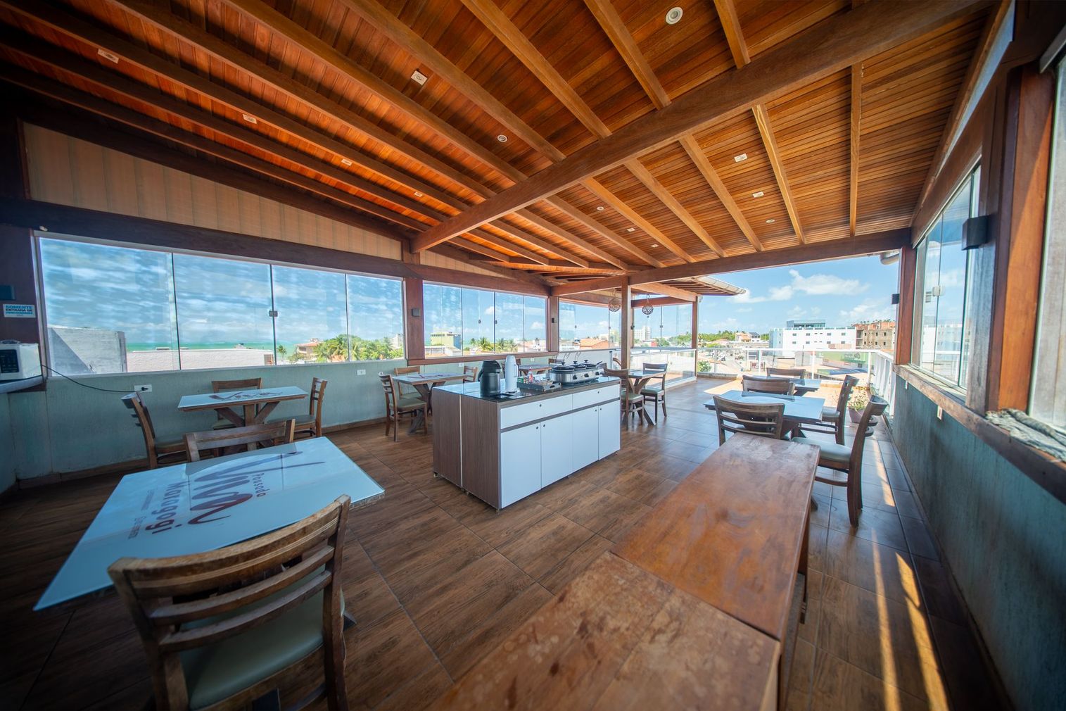 Restaurante de madeira ao ar livre com mesas, cadeiras e vista para o mar sob um teto ripado.