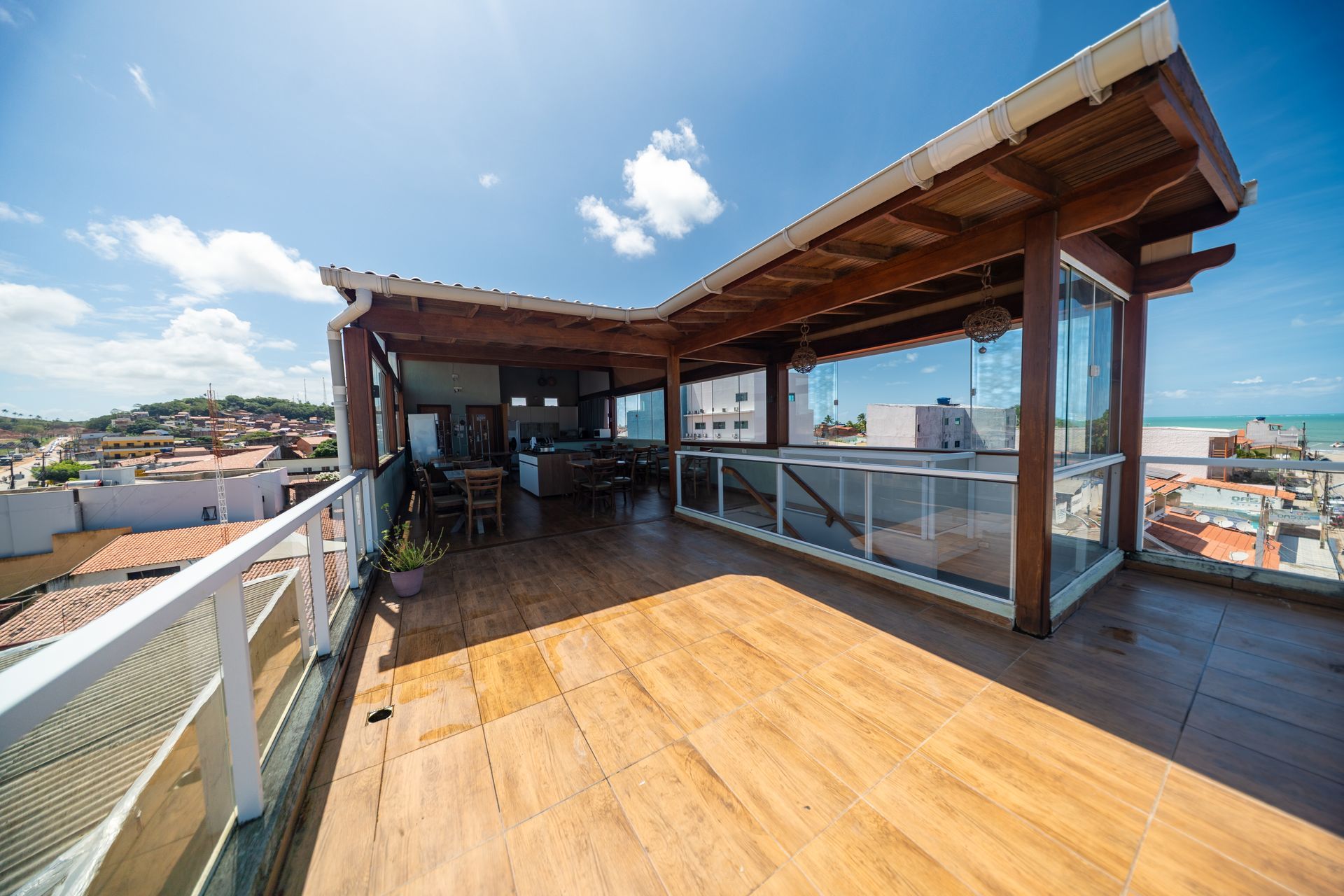 Terraço na cobertura com deck de madeira, guarda-corpo de vidro e vista para a cidade sob um céu azul.