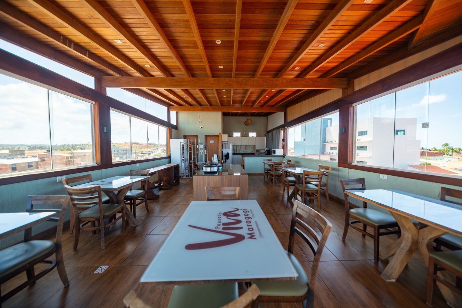 Sala de jantar iluminada com teto de madeira, mesas compridas e grandes janelas com vista para a cidade.