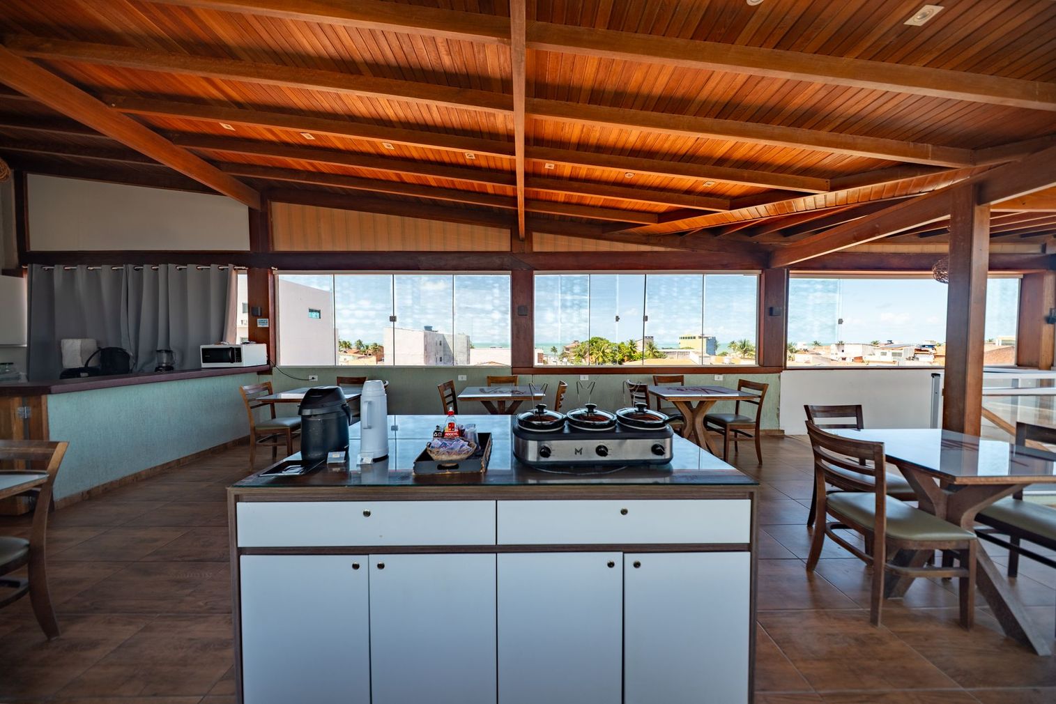Cozinha e sala de jantar integradas ao ar livre, com teto de madeira, ilha branca e vista para o mar através de grandes janelas.