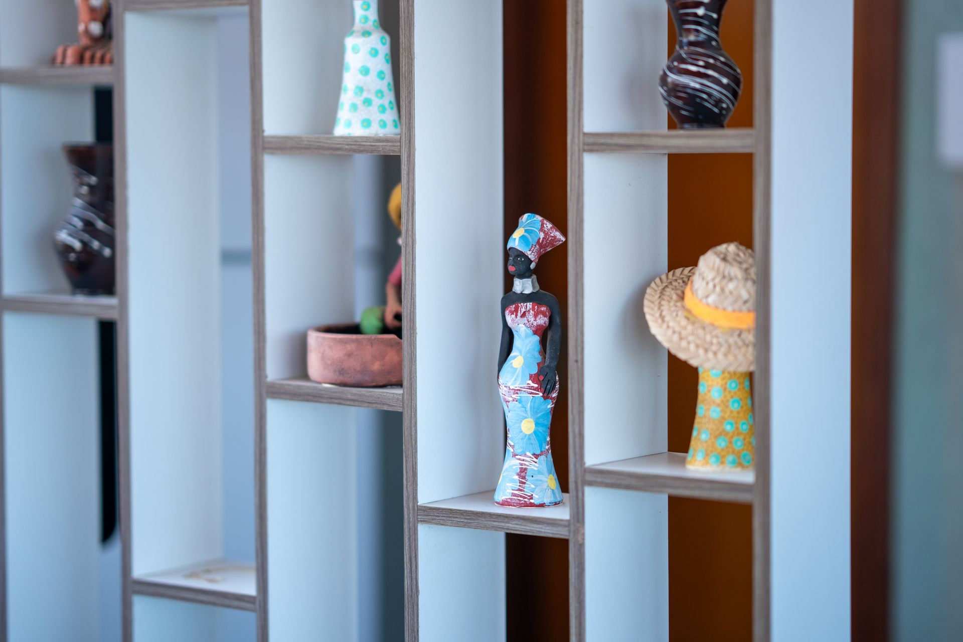 Estatuetas decorativas e chapéus expostos em estreitas prateleiras de madeira contra uma parede clara.