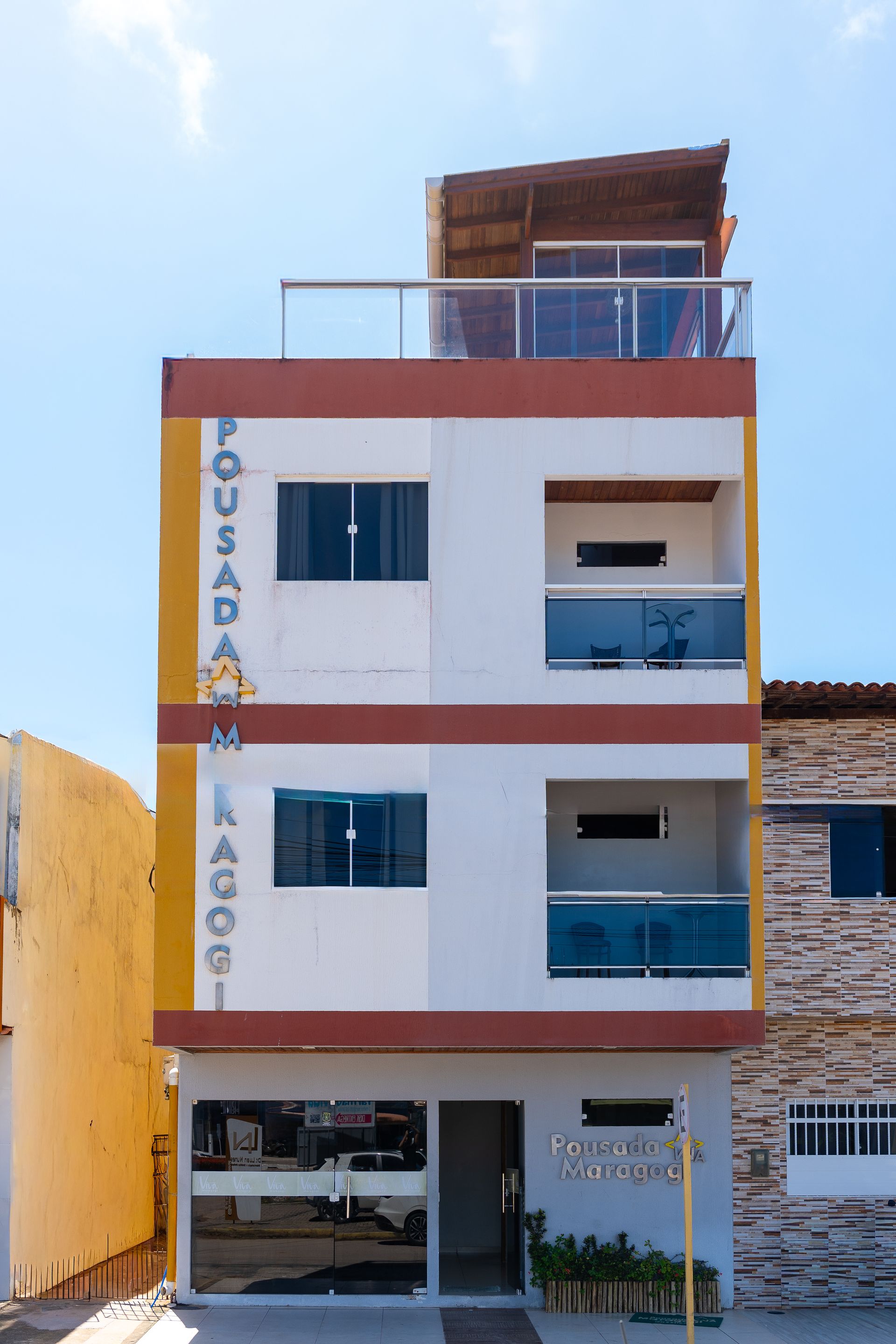 Prédio de cinco andares, branco e vermelho, com varandas e terraço na cobertura, sob um céu azul.