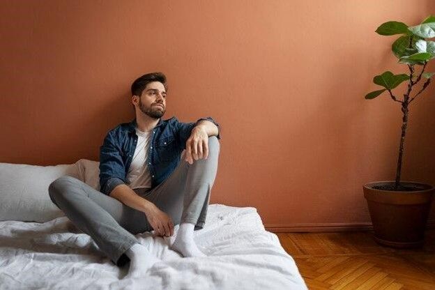 Man sits on bed, looking away, with rust-colored wall and potted plant beside him.