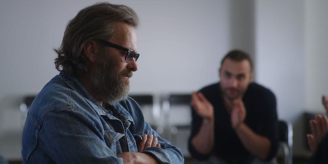 Man in denim jacket with beard, in a support group. Another man clapping in the background.