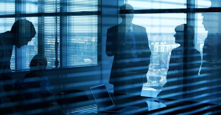 Business people silhouetted in a modern office, meeting by a window with blinds, in a blue-toned image.