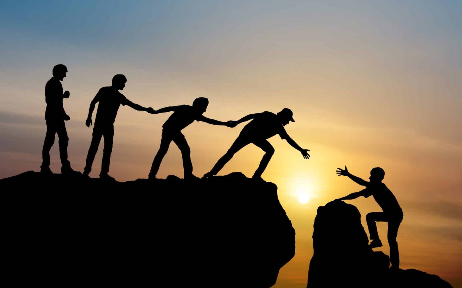 Silhouetted people helping each other climb a cliff, with a sunset in the background.