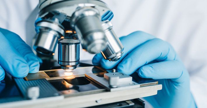 Person wearing blue gloves uses a microscope to examine a slide in a lab setting.