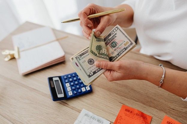 Person counting US dollar bills with a pen, calculator, and receipts on a desk.