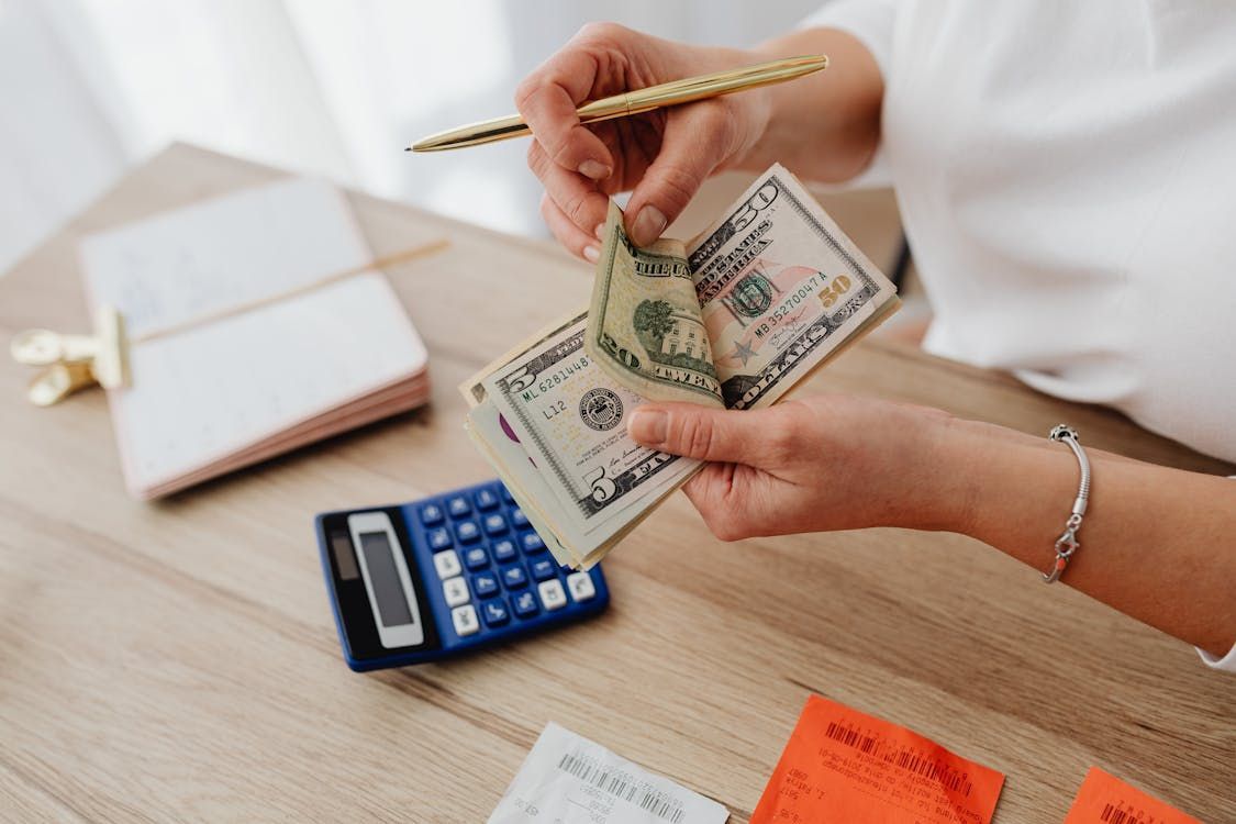 Person counting money with a calculator and notepad on a wooden desk.
