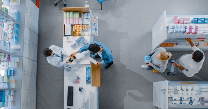 Overhead view of a pharmacy. Pharmacist and customer at the counter; others browse shelves.