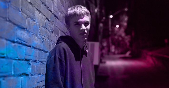Young person in a hooded sweatshirt leans against a brick wall, illuminated by blue and pink light on a city street at night.