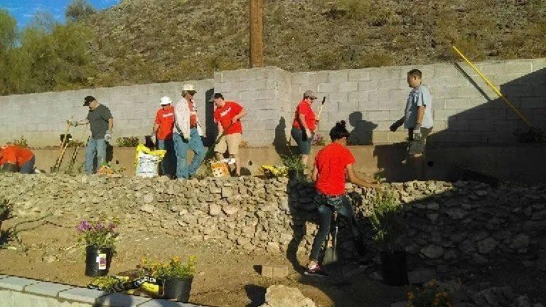 Volunteers in red shirts planting flowers and working outdoors in a sunny setting.