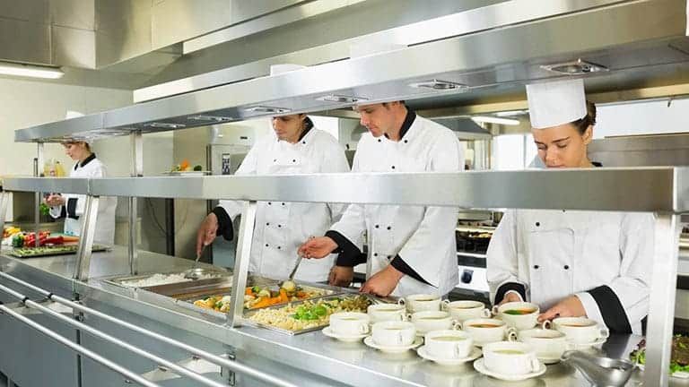 Chefs preparing food in a bright commercial kitchen. They are wearing white uniforms.