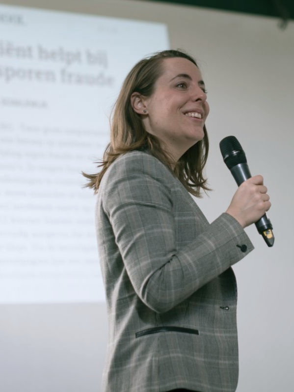 Vrouw in een grijze blazer spreekt in een microfoon voor een geprojecteerd presentatiescherm.