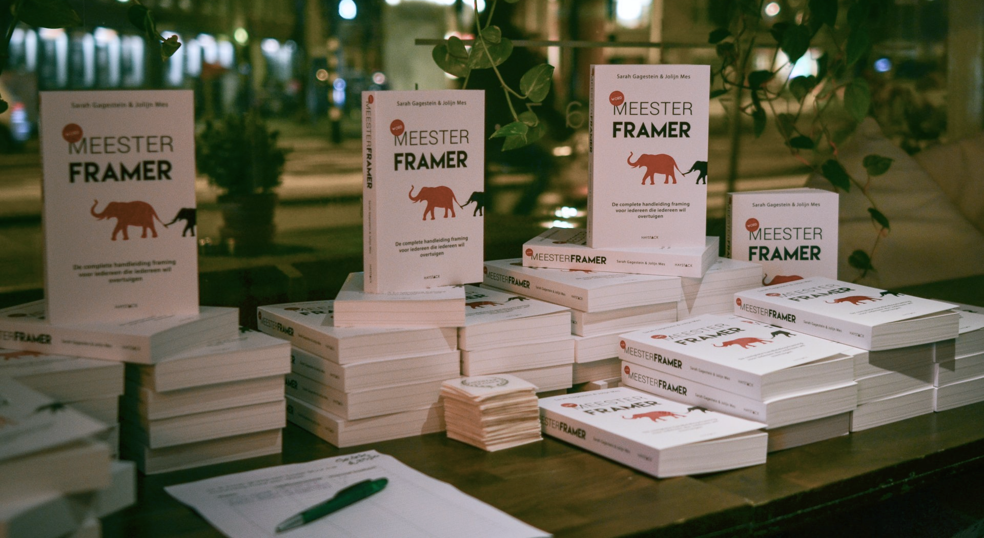 Stapels wite boeken uitgestald op een tafel, met rechtopstaande bordjes waarop 'Word Meesterframer' staat, in een boekwinkel.