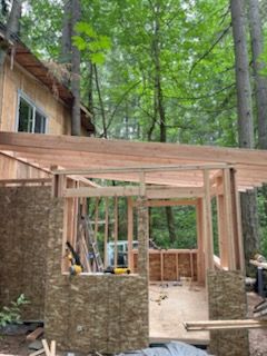 Construction of a wood-framed building addition with plywood siding, seen against a backdrop of trees.