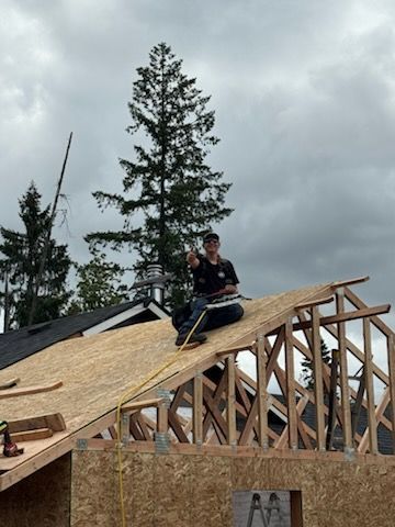 A person sits on the roof of a house under construction, pointing toward the camera with a cloudy sky in the background.