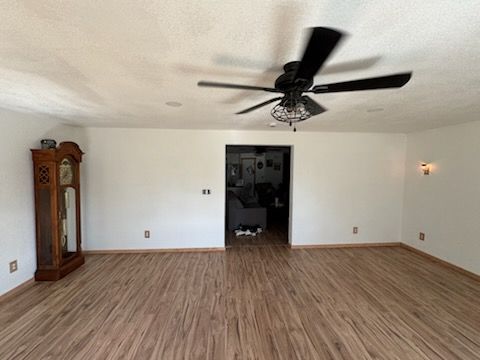 A spacious room with light wood flooring, white walls, a dark wood grandfather clock, and a black ceiling fan.