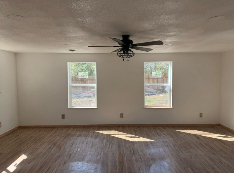 An empty room with white walls, light wood flooring, two windows, and a dark ceiling fan centered overhead.