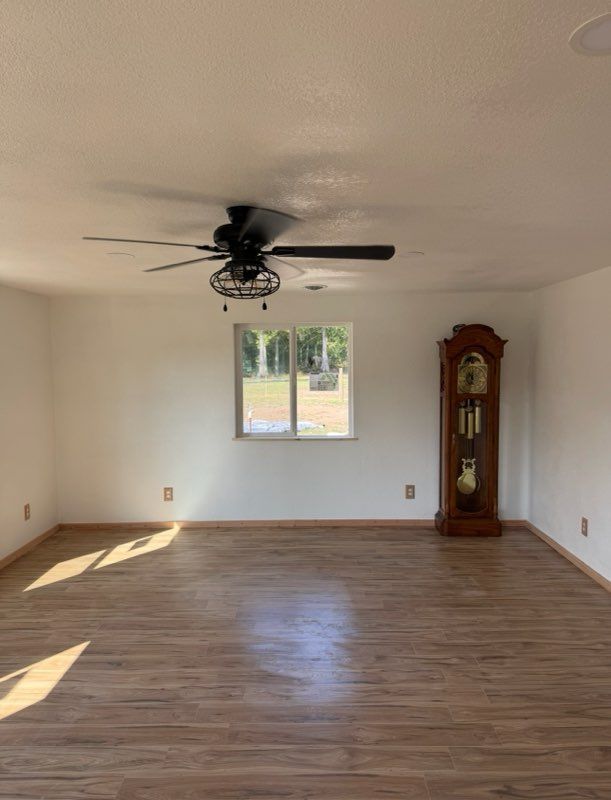 A spacious room with light wood flooring, white walls, a ceiling fan, a centered window, and a tall wooden grandfather clock.