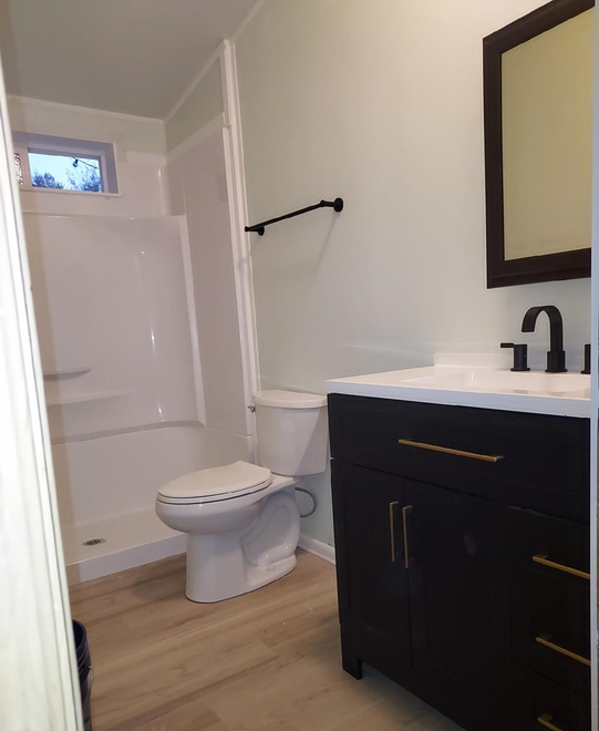 A modern bathroom with light walls, a wood-look floor, a white shower stall, a toilet, and a dark vanity with a mirror.