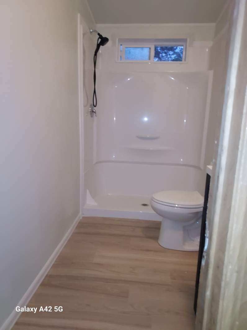 A small, bright bathroom with light wood flooring, a white shower stall with a black handheld showerhead, and a toilet.