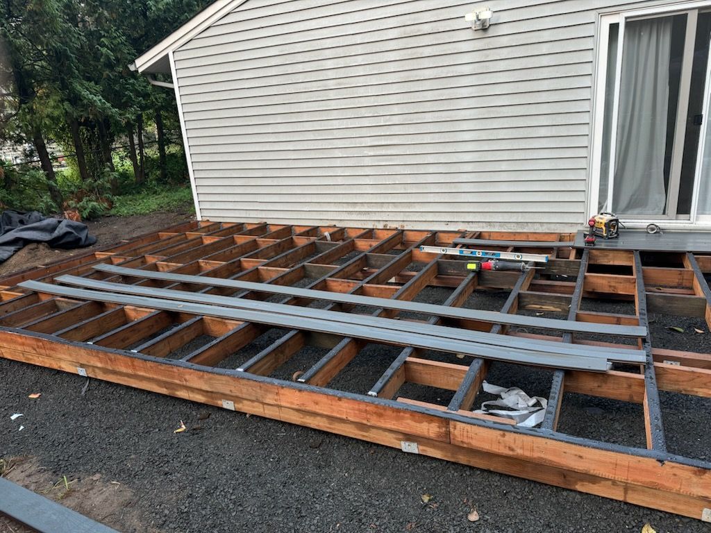 An unfinished wooden deck frame under construction against a house with light grey vinyl siding.