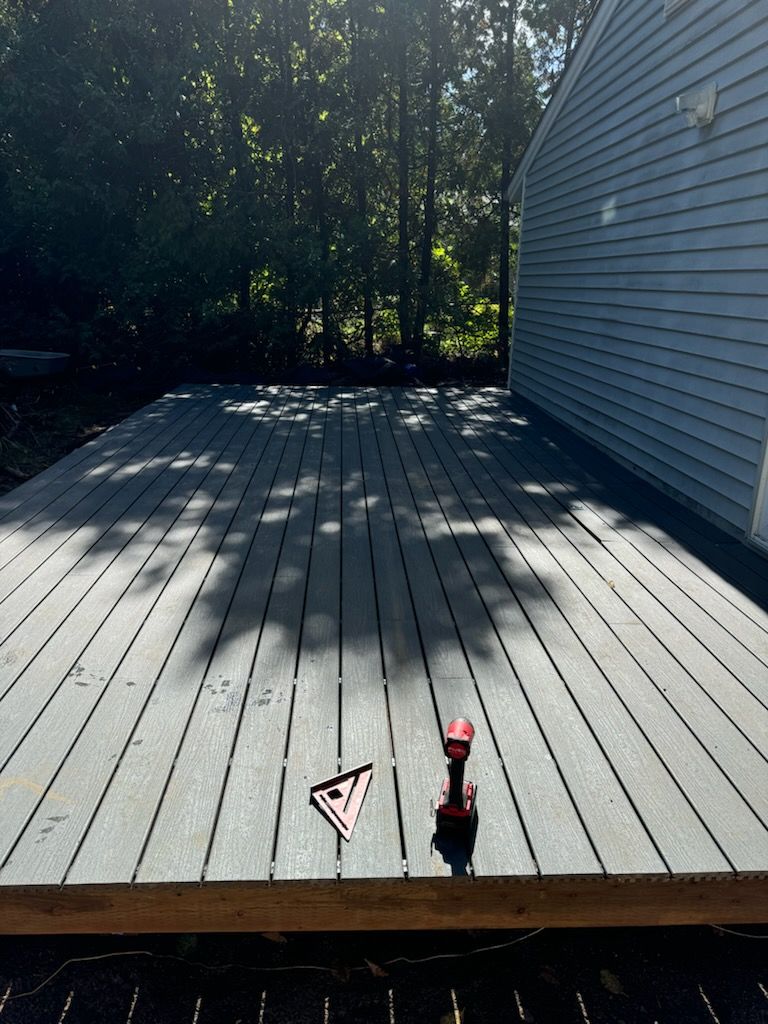 A gray composite deck under construction attached to a house, with a speed square and cordless drill on the surface.