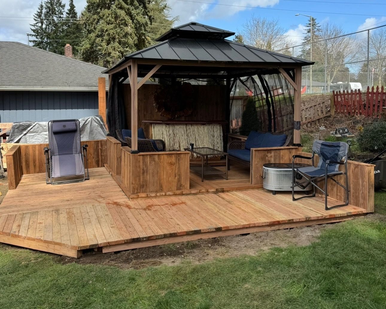 A backyard wooden deck features a gazebo covering a lounge area, with outdoor chairs and a small table on the wood surface.