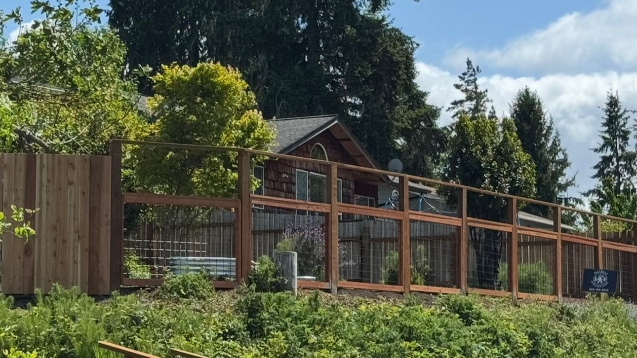 A wooden fence surrounds a rustic house tucked among green trees under a partly cloudy blue sky.