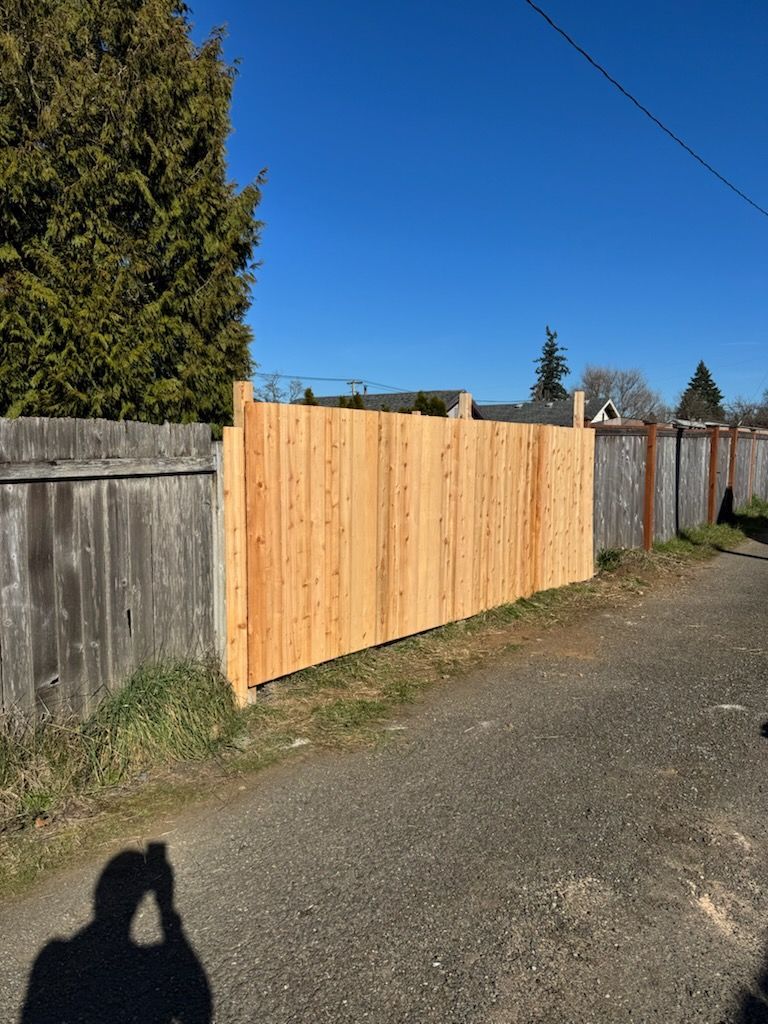 A new, light-colored wooden fence section stands between two older, weathered grey fences along a gravel path.