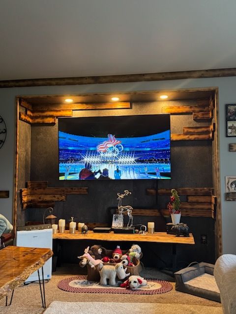 A TV mounted on a dark wall with rustic wood accents and overhead lights, above a wood shelf with decor and plush toys.