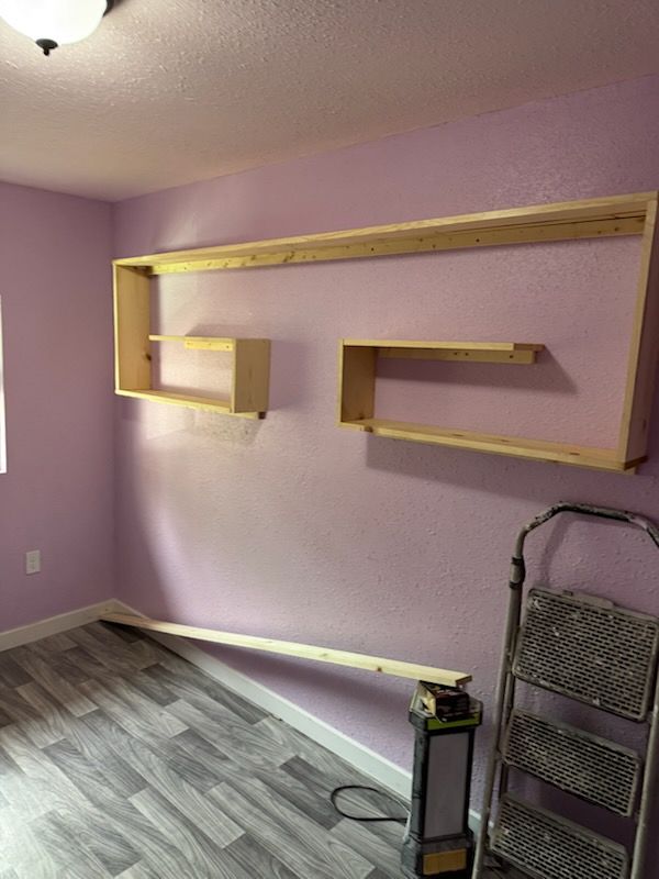 A wooden shelf frame installed on a light purple wall, with a loose board leaning on the floor and a metal step ladder.