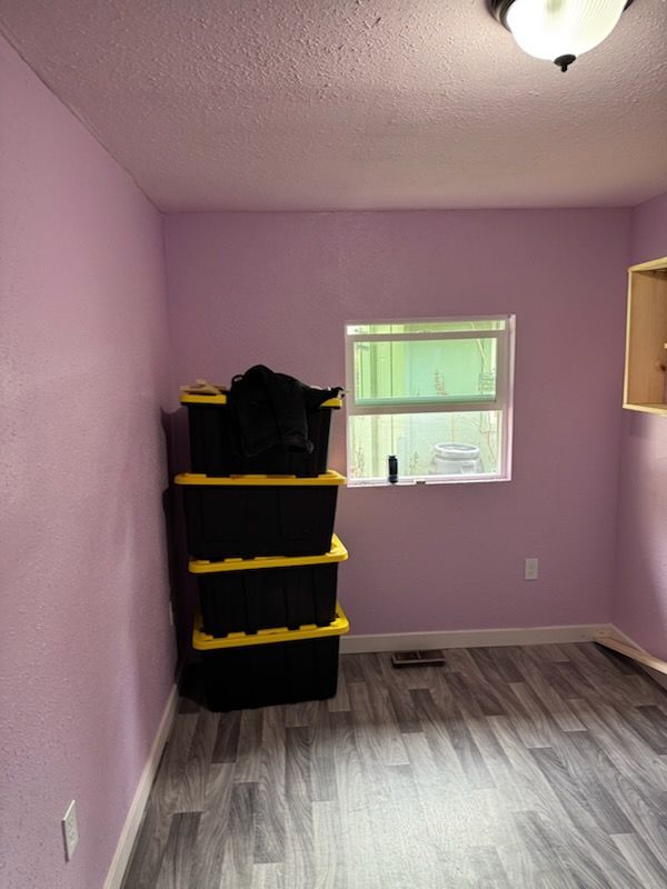 A stack of four black storage bins with yellow lids sits in the corner of a room with light purple walls and gray flooring.