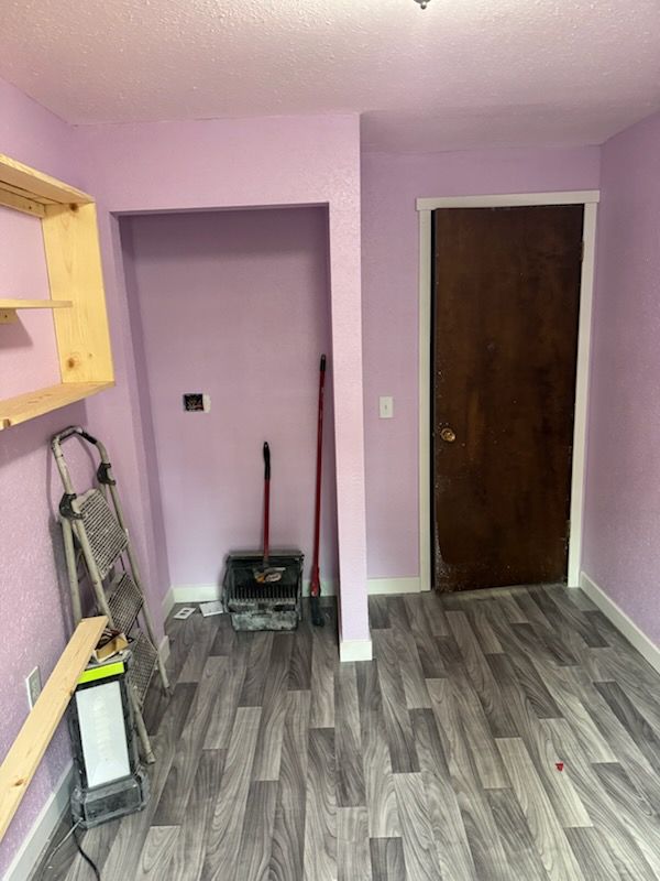 A small room with light purple walls, grey wood-look flooring, a wooden shelf, a step ladder, and a dark wooden door.