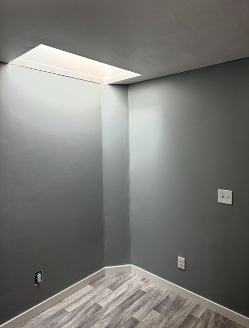 An empty corner of a room with grey walls, white baseboards, light-colored wood flooring, and a rectangular skylight.