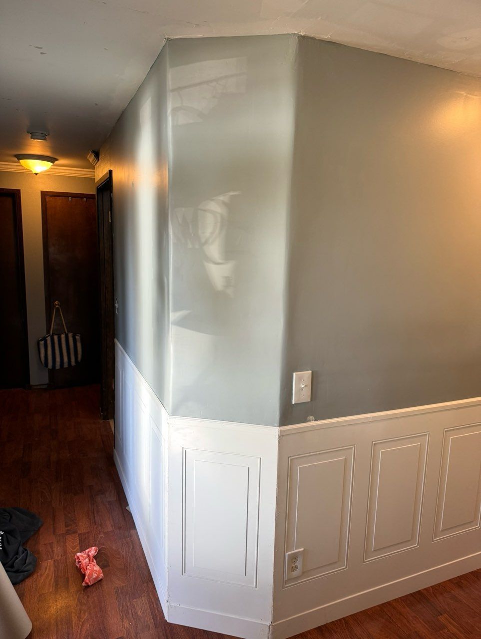 An interior hallway with grey walls above white wainscoting and wooden flooring, featuring a corner with a light switch.