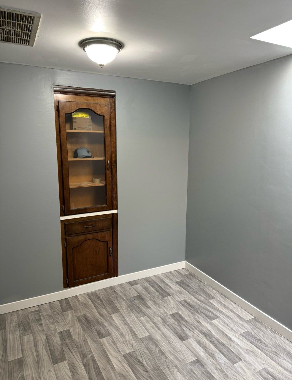 A room with gray walls, light-toned wood-look plank flooring, a ceiling light, and a built-in wooden cabinet.