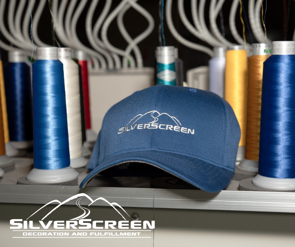 Blue SilverScreen cap centered among thread spools in a sewing workshop with SilverScreen logo watermark