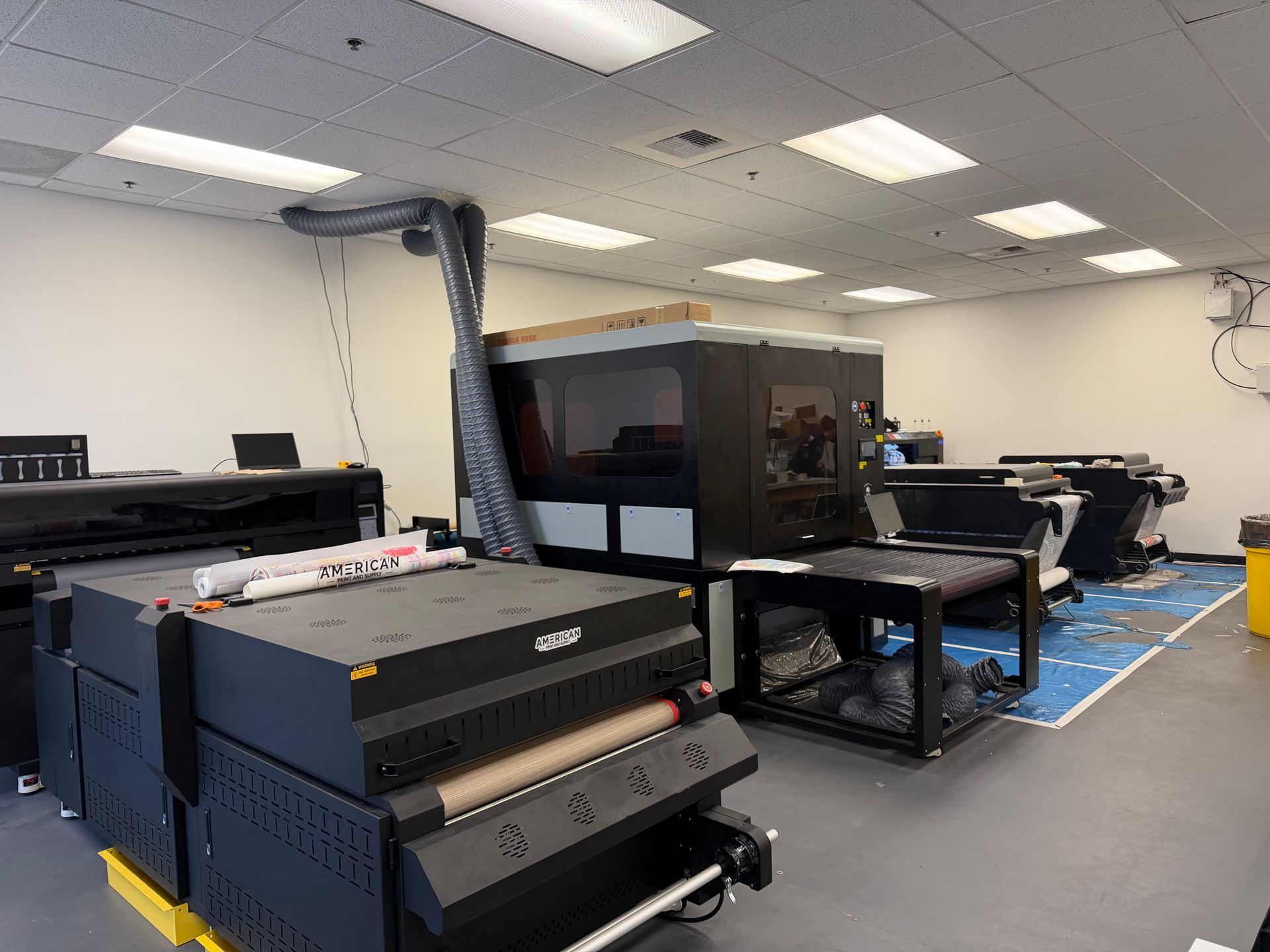 A well-lit room featuring several large industrial digital printing machines and specialized equipment on a gray floor.