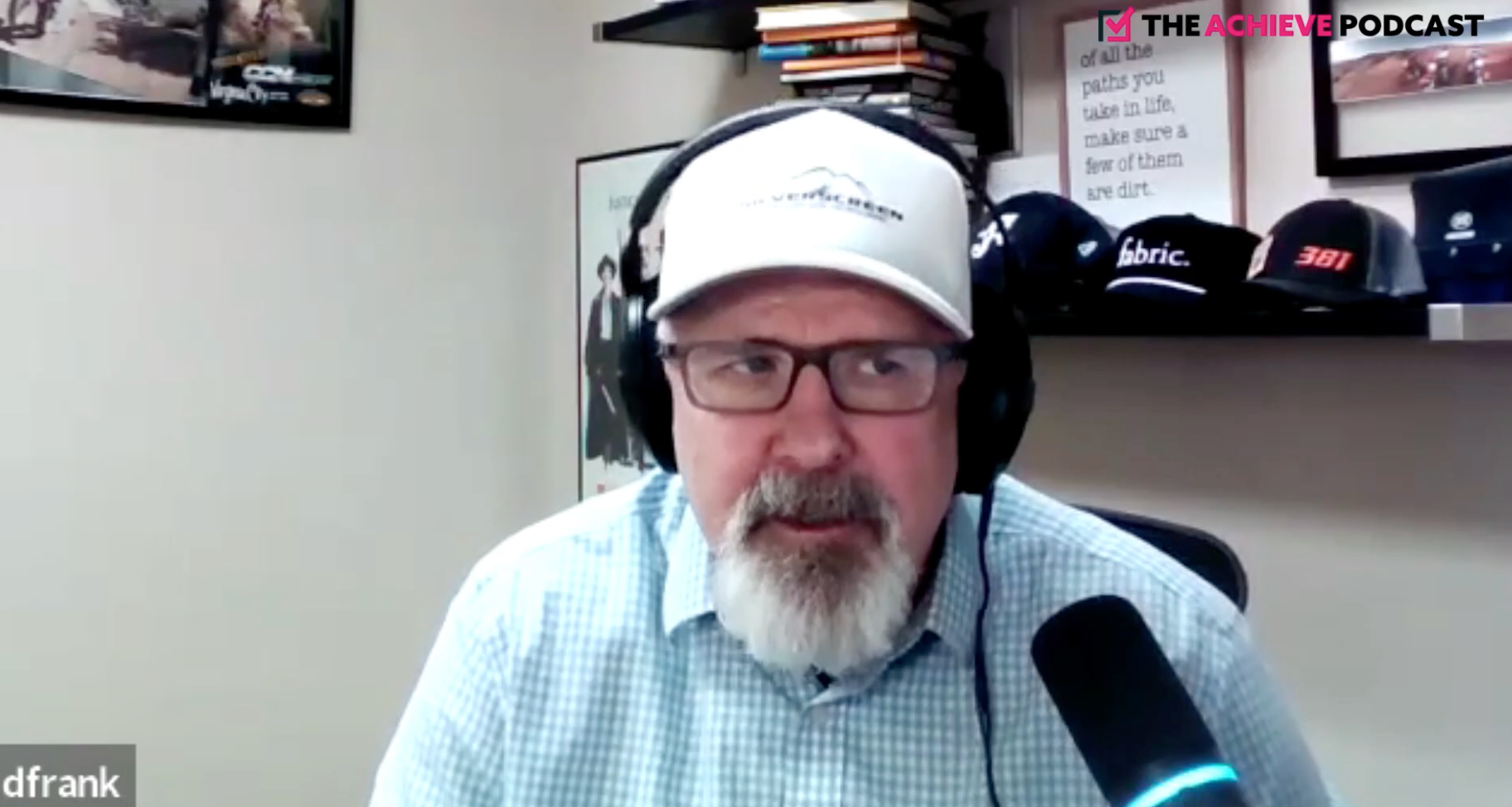 Podcast host wearing a white cap and glasses speaks into a microphone in a home office.