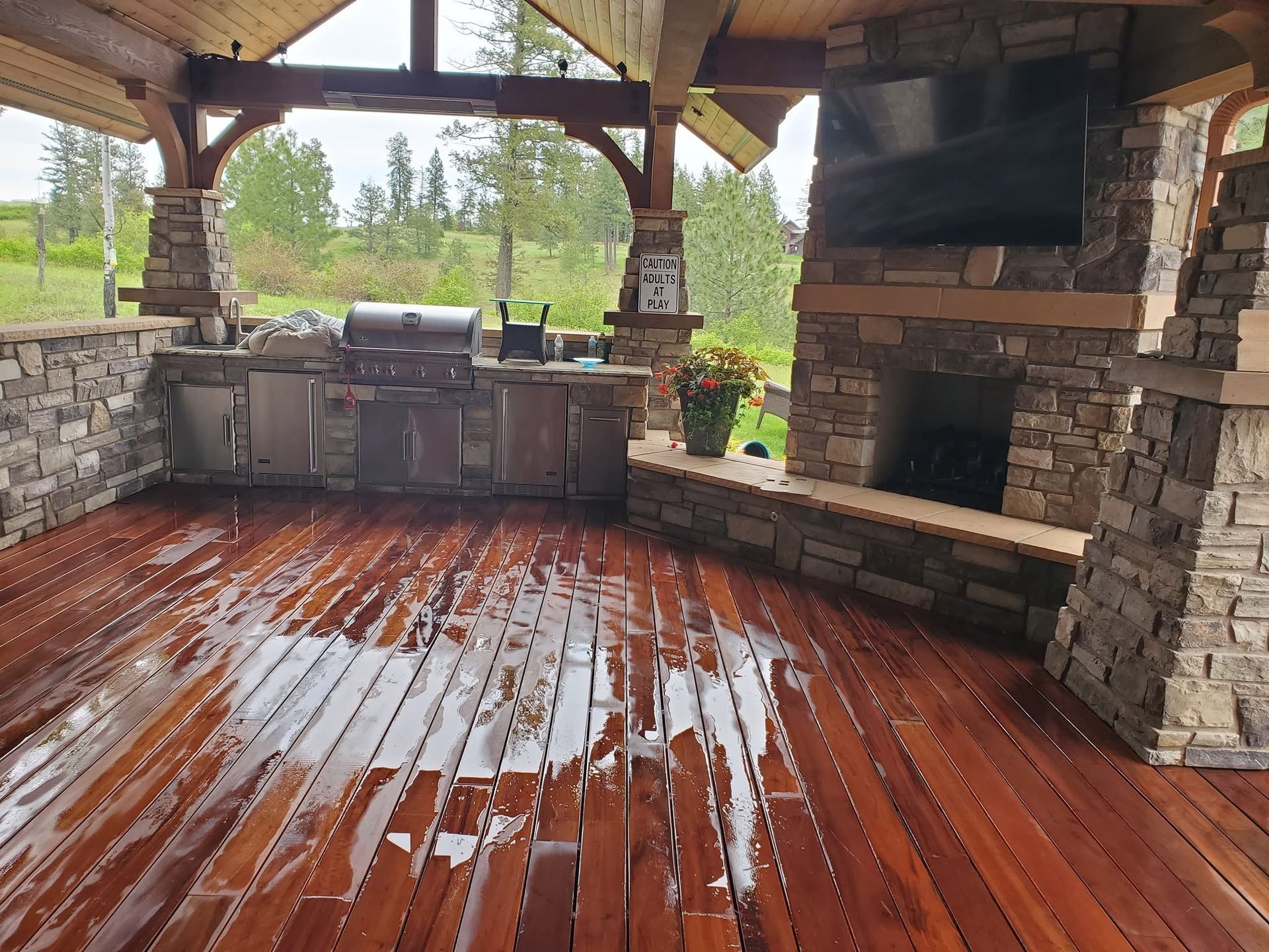 Covered outdoor kitchen with stone fireplace and wet wooden floor overlooking greenery