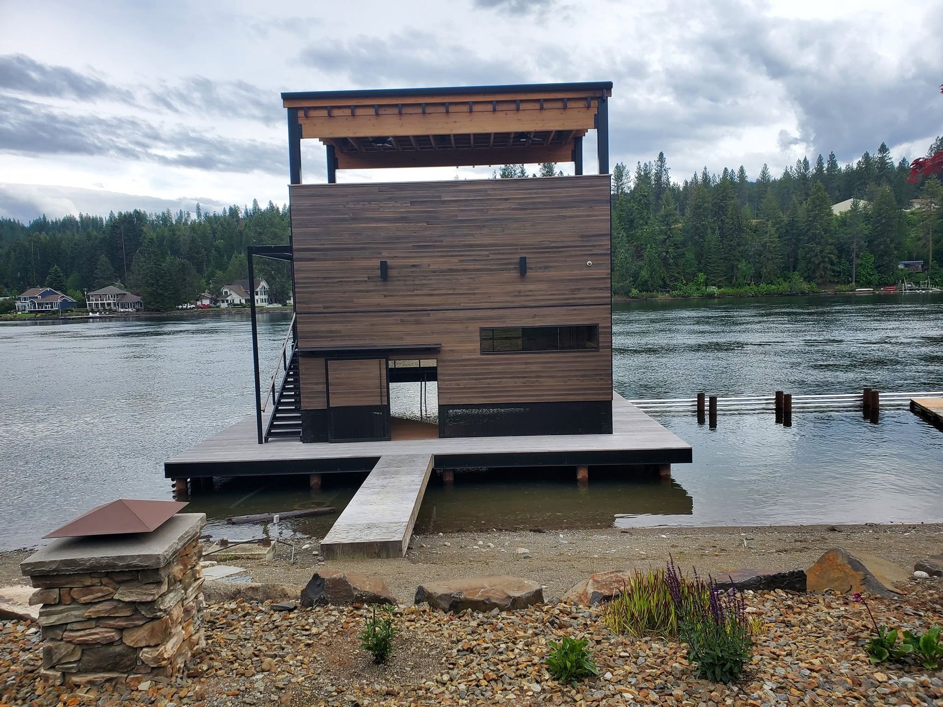 Floating wooden cabin on a lake shore with a dock, under cloudy skies, surrounded by forested hills